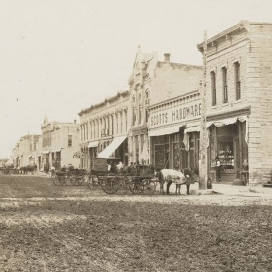 Historic street scene featuring horse-drawn carriages and storefronts, including a hardware shop.