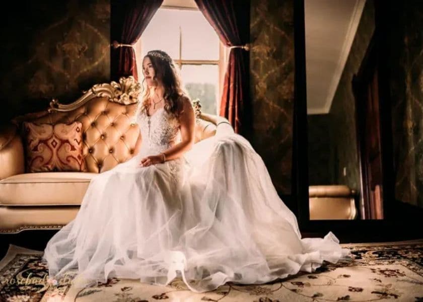 A bride in a flowing wedding dress sits thoughtfully on a vintage sofa, with a large mirror reflecting the elegant setting. A bride in a flowing wedding dress sits thoughtfully on a vintage sofa, with a large mirror reflecting the elegant setting.