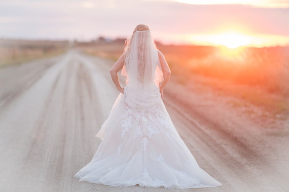 A bride stands on a dirt road at sunset, gazing into the distance.