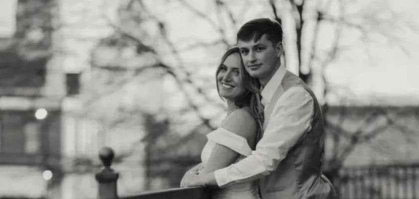 A couple embraces on a balcony, smiling against a blurred backdrop of trees.