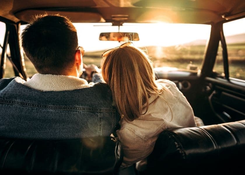 A couple embraces in a vintage car, with the sun setting in the background.