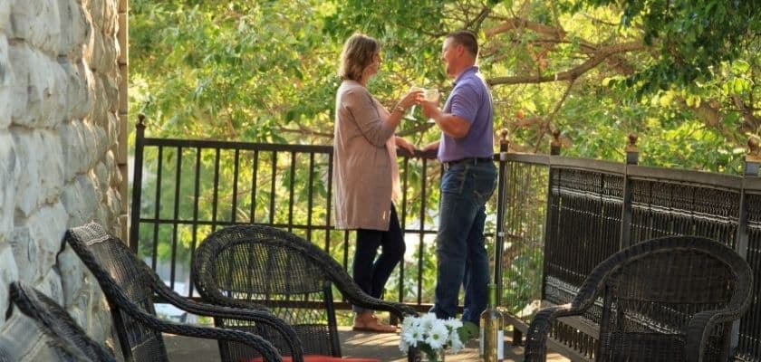 A couple stands on a balcony, smiling and holding hands amidst a backdrop of greenery.