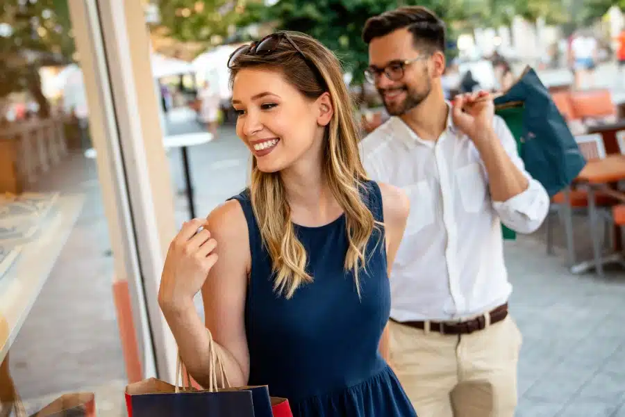 A woman smiles while looking into a shop window, accompanied by a man holding shopping bags.
