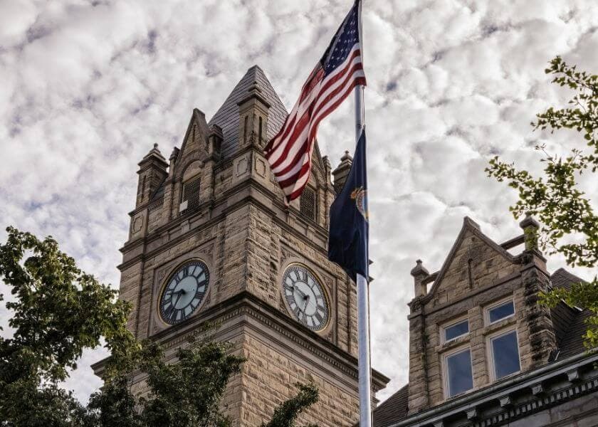 A historic clock tower with an American flag flying in front against a cloudy sky.
