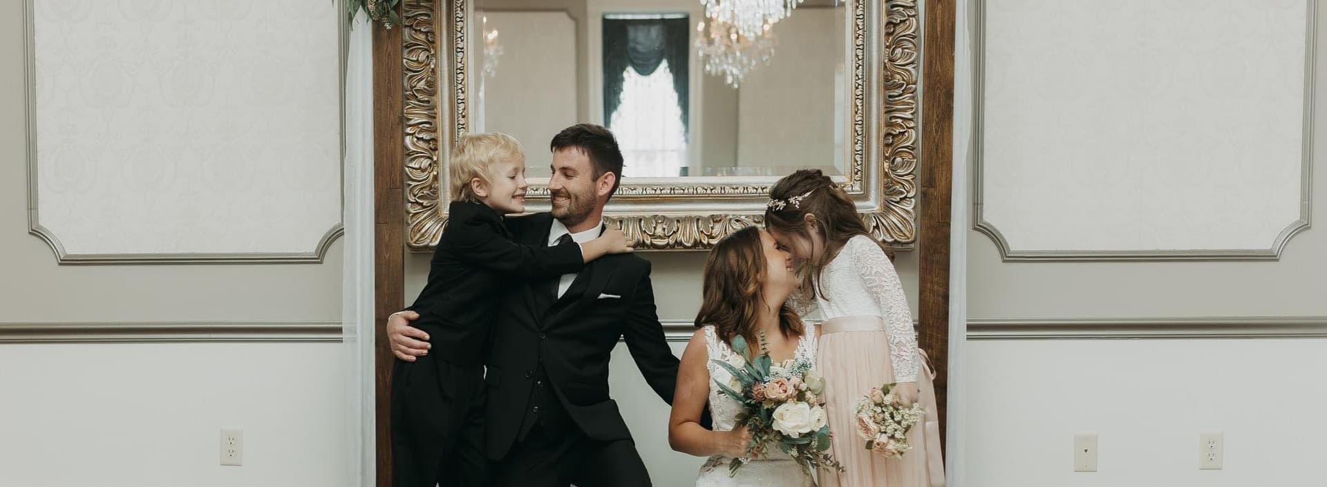A groom holds a smiling child while a bride shares a joyful moment with another young girl in a beautifully decorated room.