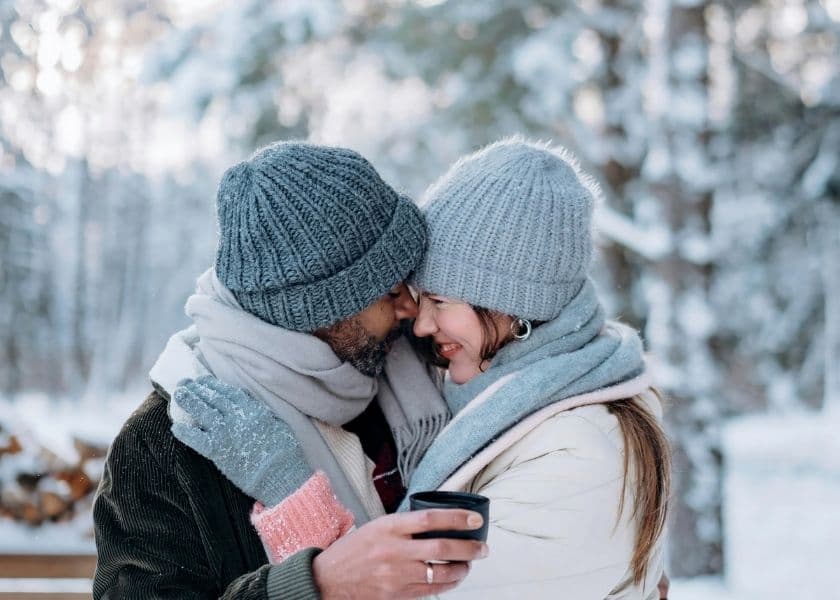 A couple embraces in a snowy setting, wearing cozy winter hats and scarves.