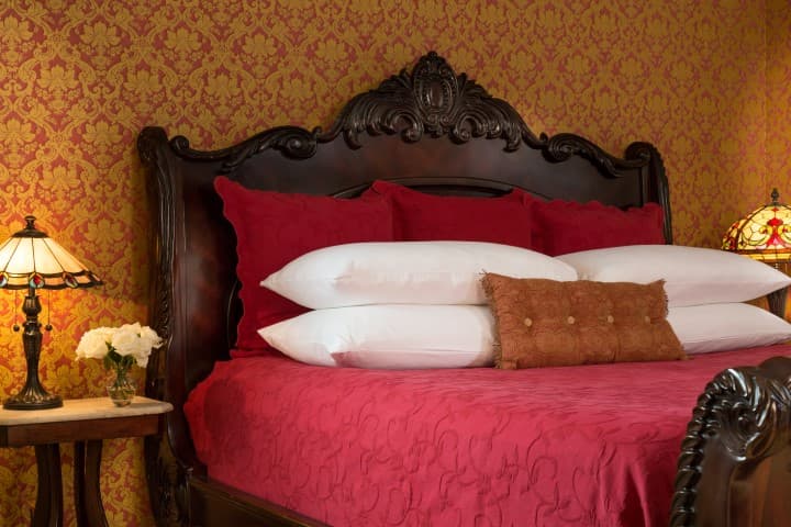 A luxurious bedroom featuring an ornate wooden bed with red bedding and decorative pillows, alongside antique lamps and a floral arrangement.