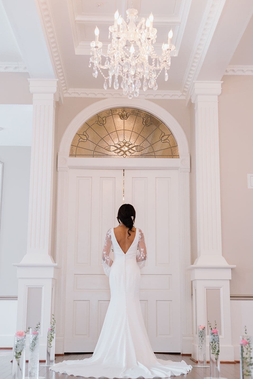 A bride in a backless wedding gown stands before elegant double doors under a chandelier.