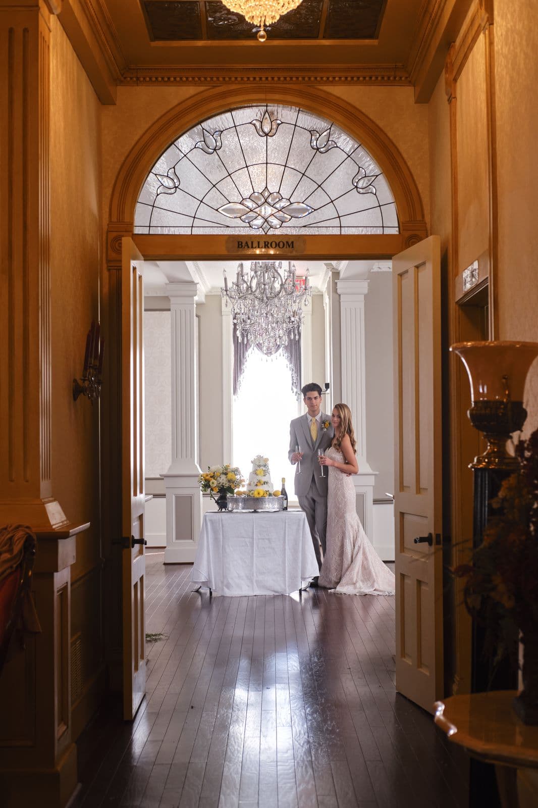 A couple stands together in a ballroom, holding flutes and smiling in front of a decorated table.