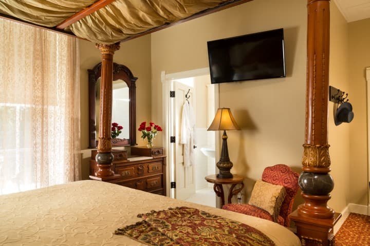 A cozy bedroom featuring a four-poster bed, a vintage dresser, a lamp, and a television, with a bathroom door partially visible.
