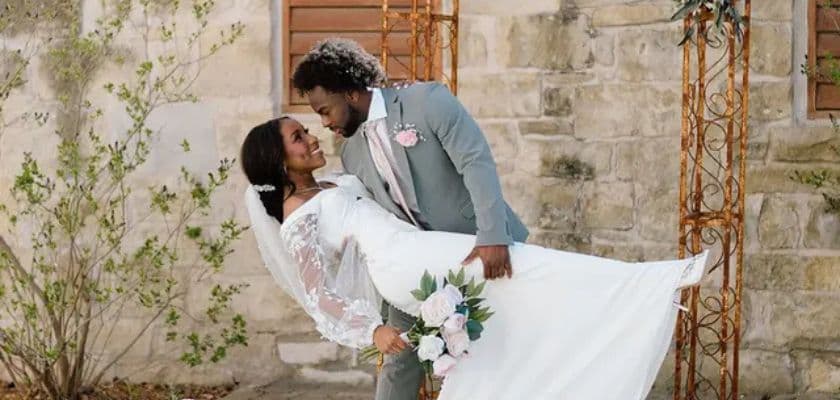 A newlywed couple shares a romantic moment outdoors, with the bride elegantly dressed in white and the groom in a gray suit.