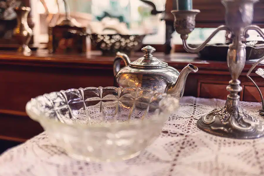 A glass bowl and a silver teapot are displayed on a lace tablecloth.