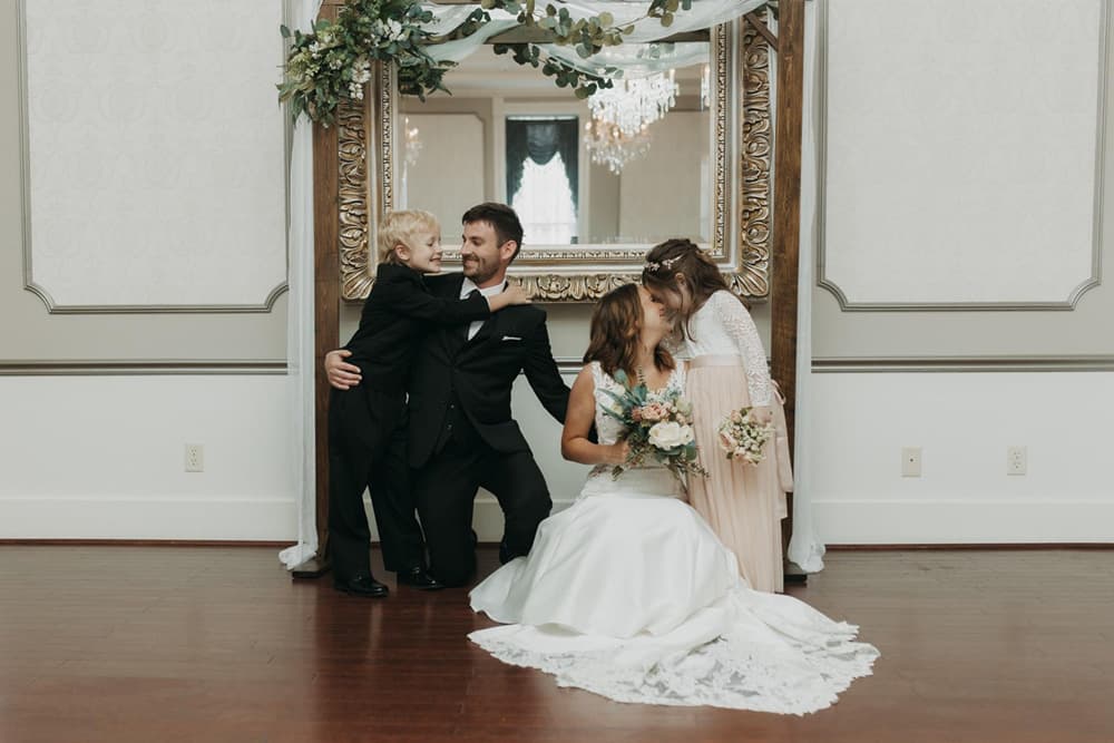 A bride and groom share a tender moment as two children embrace them in front of an ornate mirror.