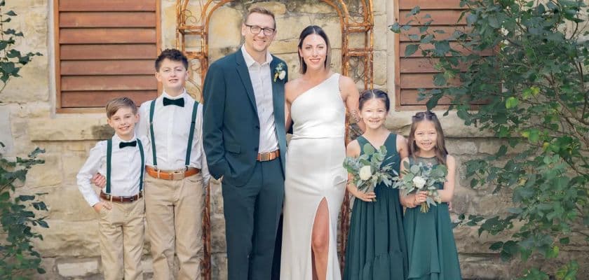 A bride in a white dress stands with a groom and four children in formal attire against a rustic backdrop.