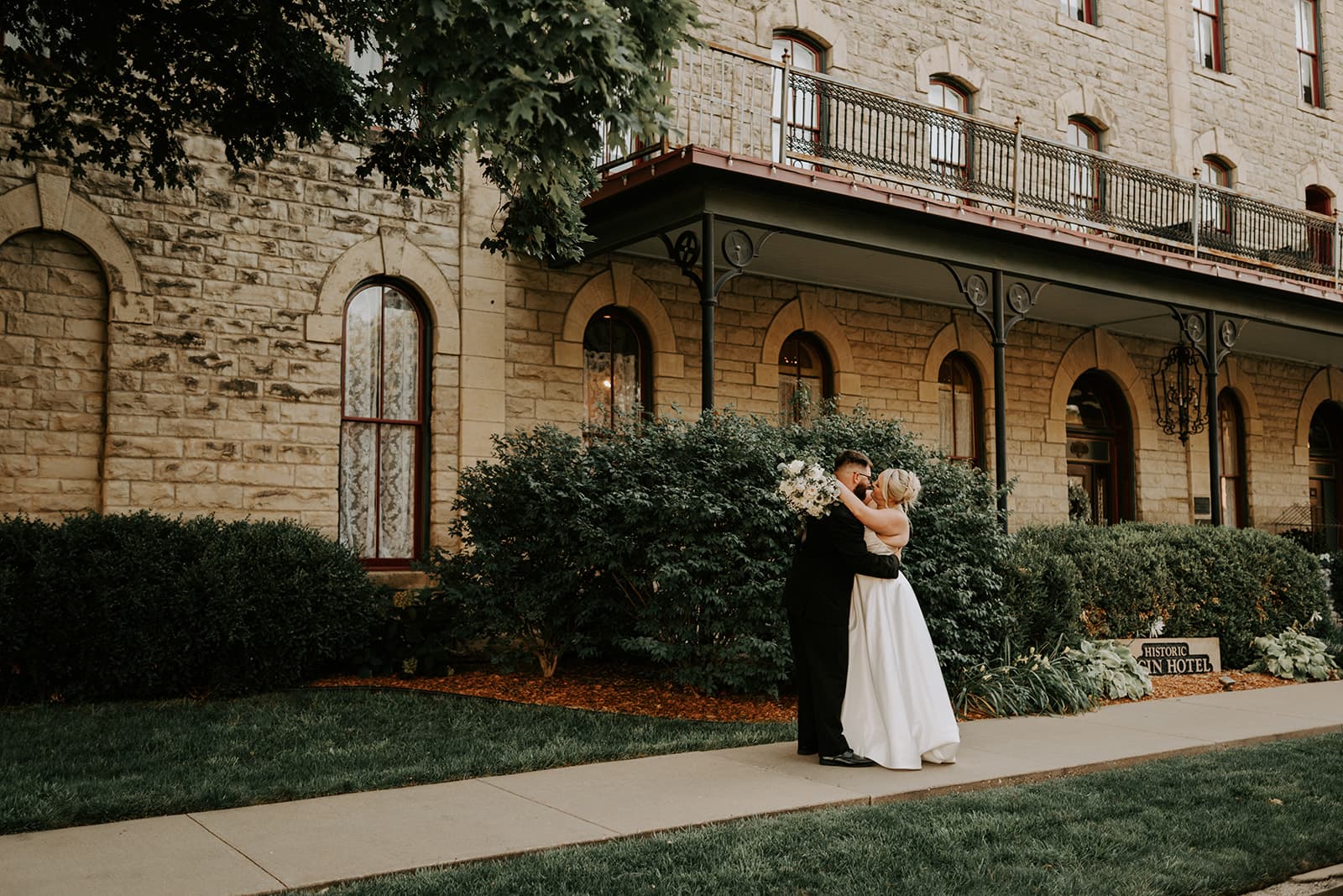 A bride and groom embrace in front of a historic stone building, surrounded by greenery.