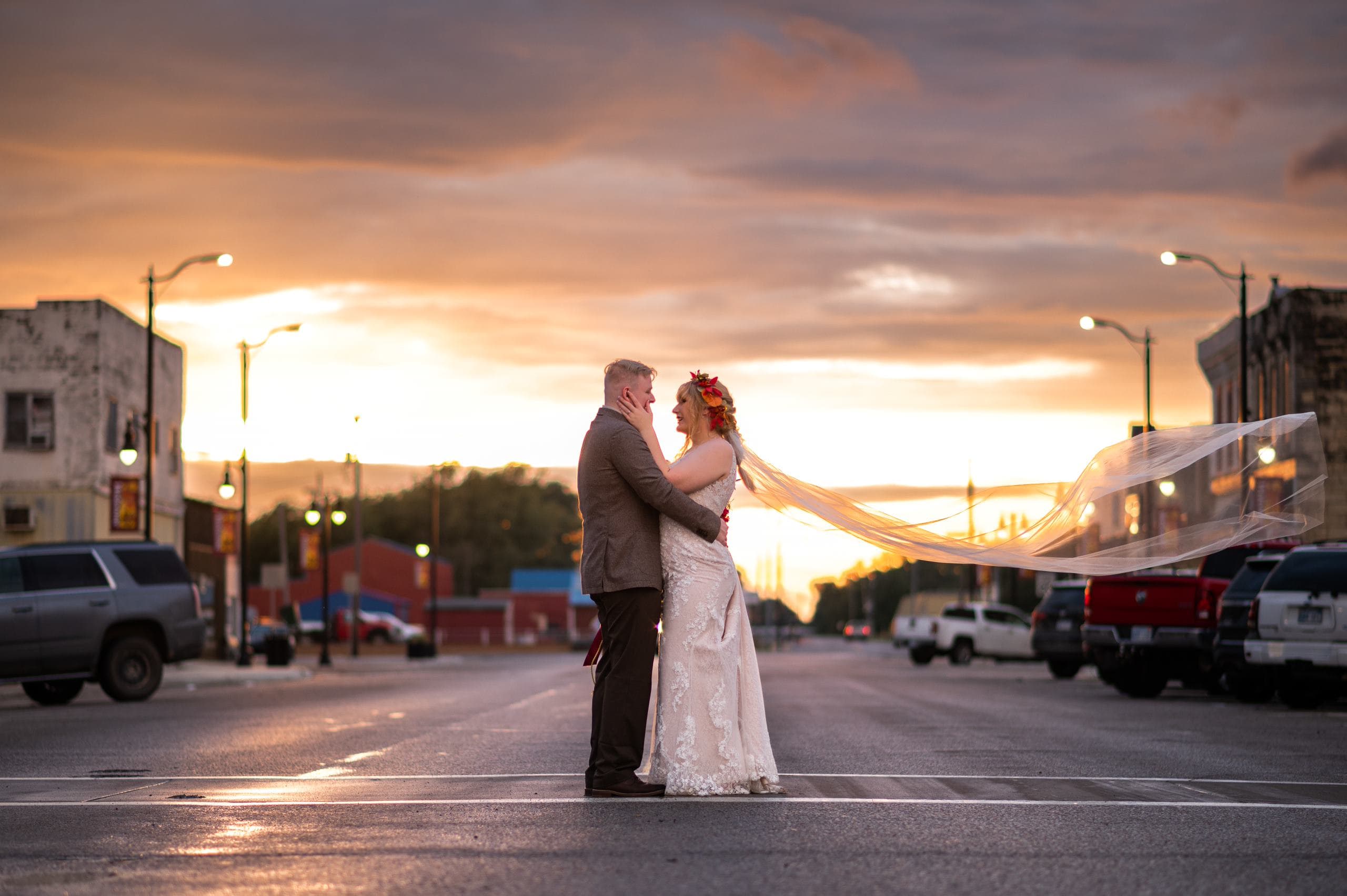A couple lovingly embraces in a deserted street at sunset, with the bride's veil flowing in the wind.