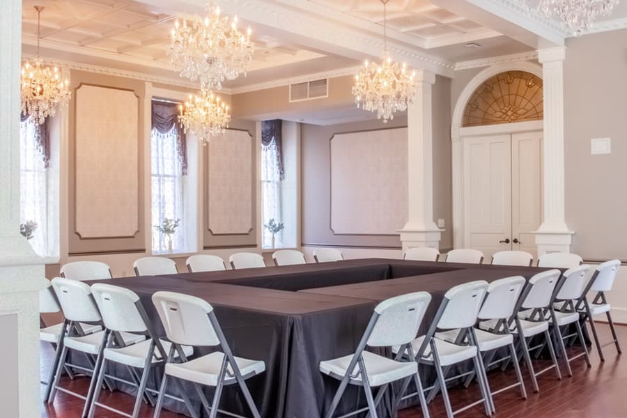 A spacious meeting room with a large black table surrounded by folding chairs, illuminated by chandeliers.