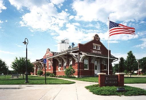 A historic brick library building with a flagpole and American flag, surrounded by greenery under a partly cloudy sky.