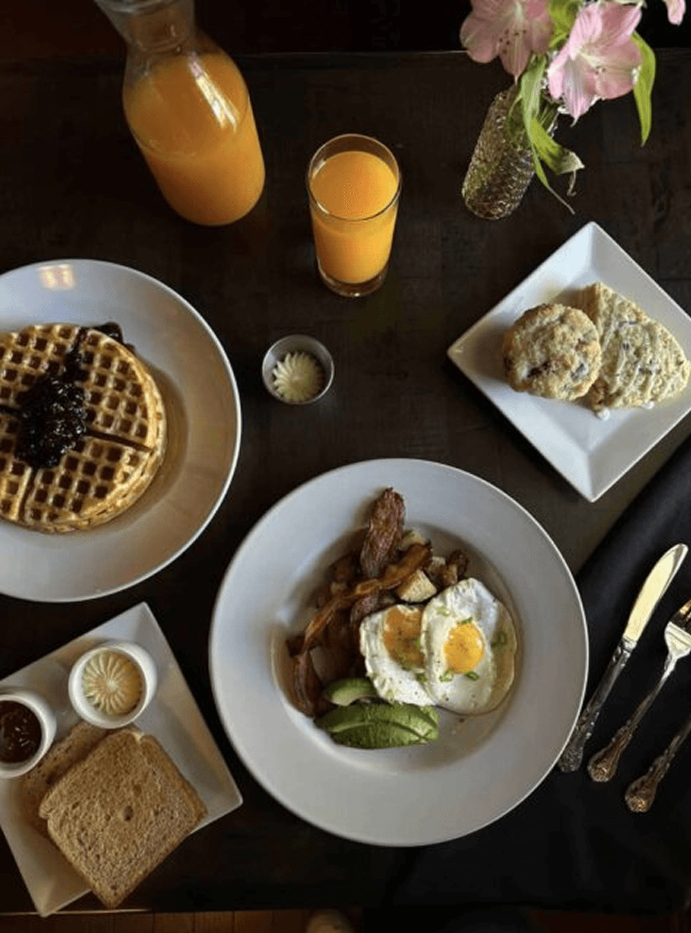 A top-down view of a breakfast spread featuring waffles, eggs, bacon, toast, cookies, and orange juice.