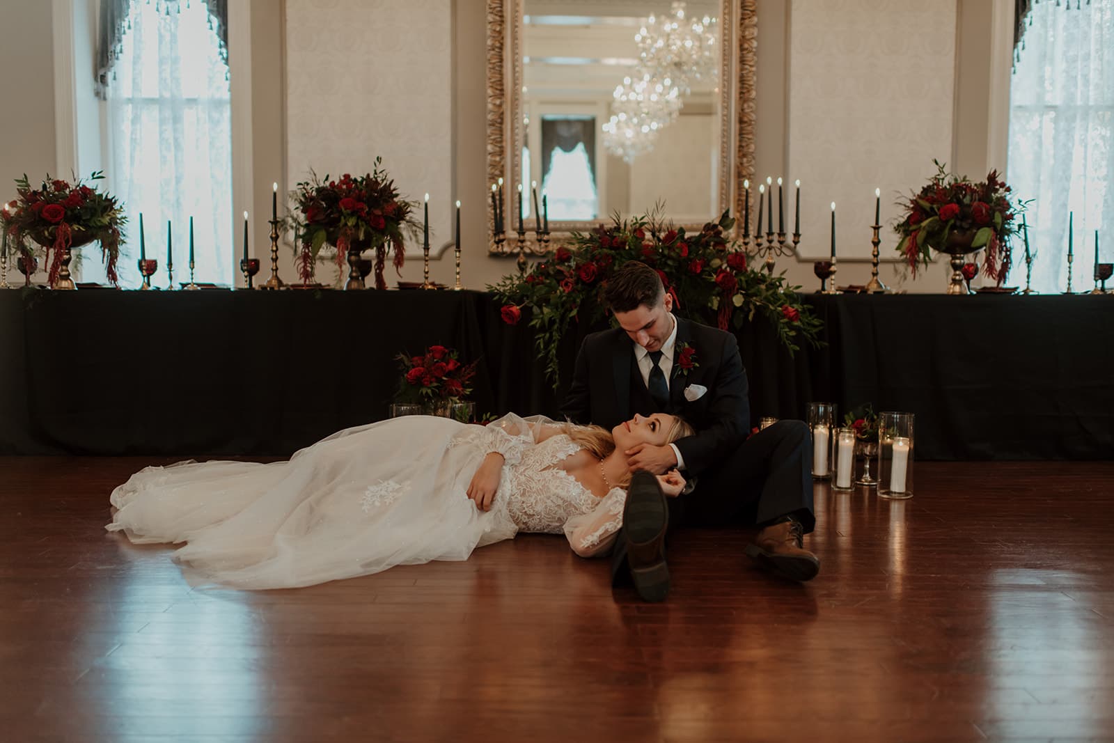 A couple in elegant attire poses on the floor surrounded by floral arrangements and candles, with a mirror reflecting the scene.