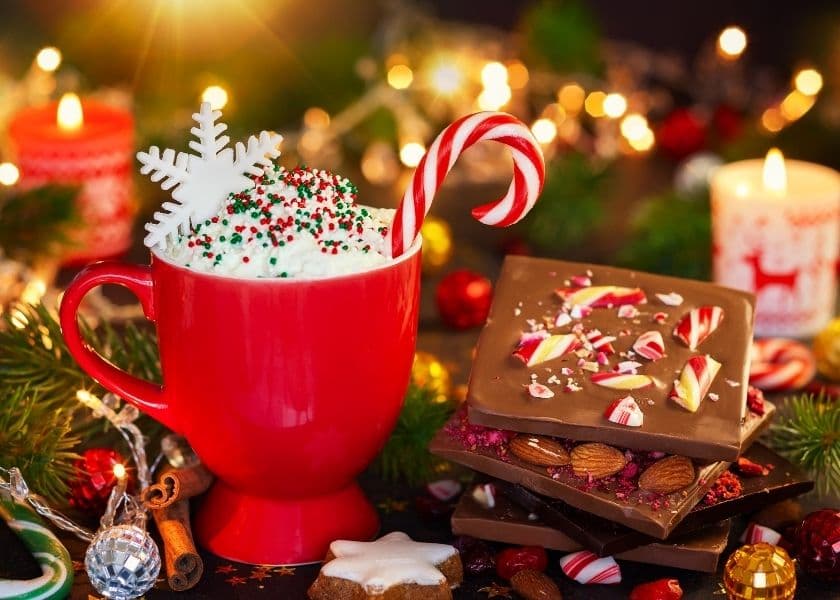 A festive red mug filled with whipped cream, topped with a snowflake and candy cane, surrounded by holiday treats and decorations.