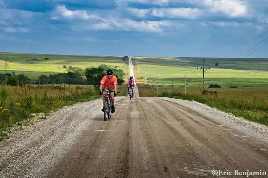 Two cyclists ride on a gravel road surrounded by expansive green fields under a cloudy sky.
