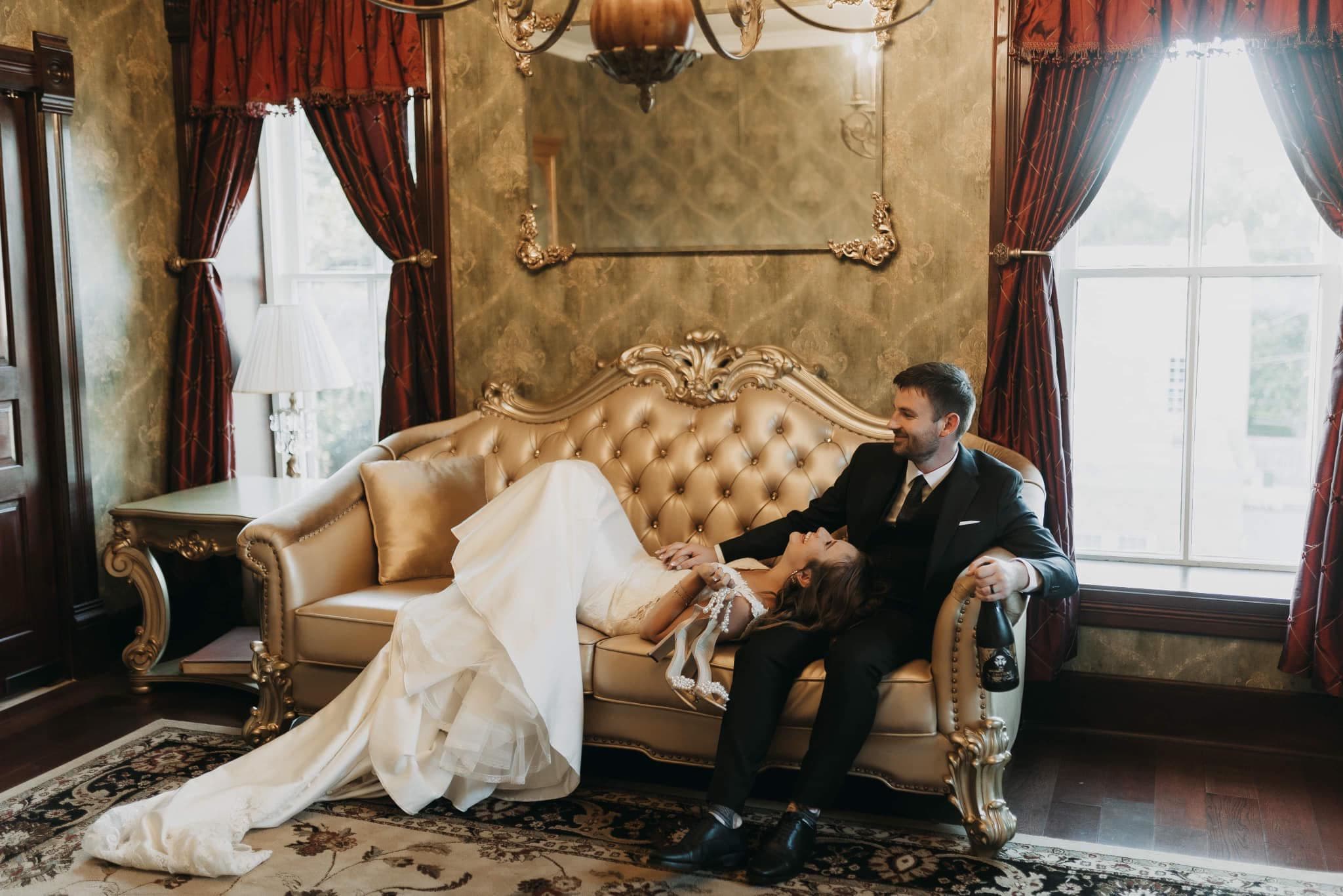 A couple sits on an elegant sofa, with the bride playfully reclining and the groom smiling beside her.