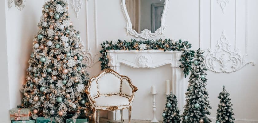 A beautifully decorated Christmas tree stands next to a white mantel, surrounded by smaller evergreen trees and presents.