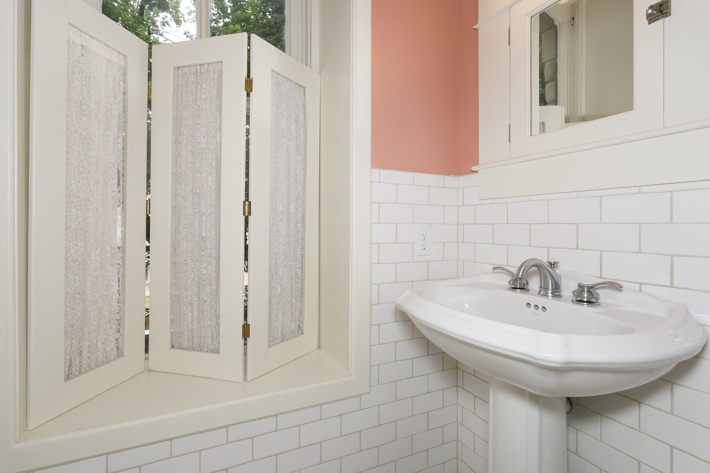 A white pedestal sink is positioned against a tiled wall with a window featuring decorative shutters above it.