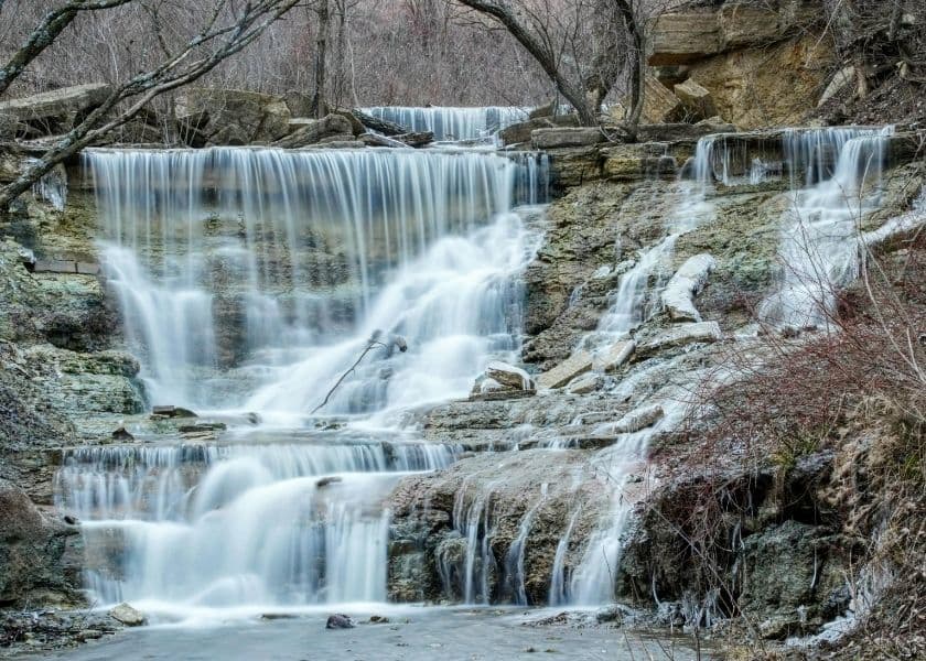 A serene waterfall cascades over rocky layers surrounded by bare trees and muted vegetation. A serene waterfall cascades over rocky layers surrounded by bare trees and muted vegetation.