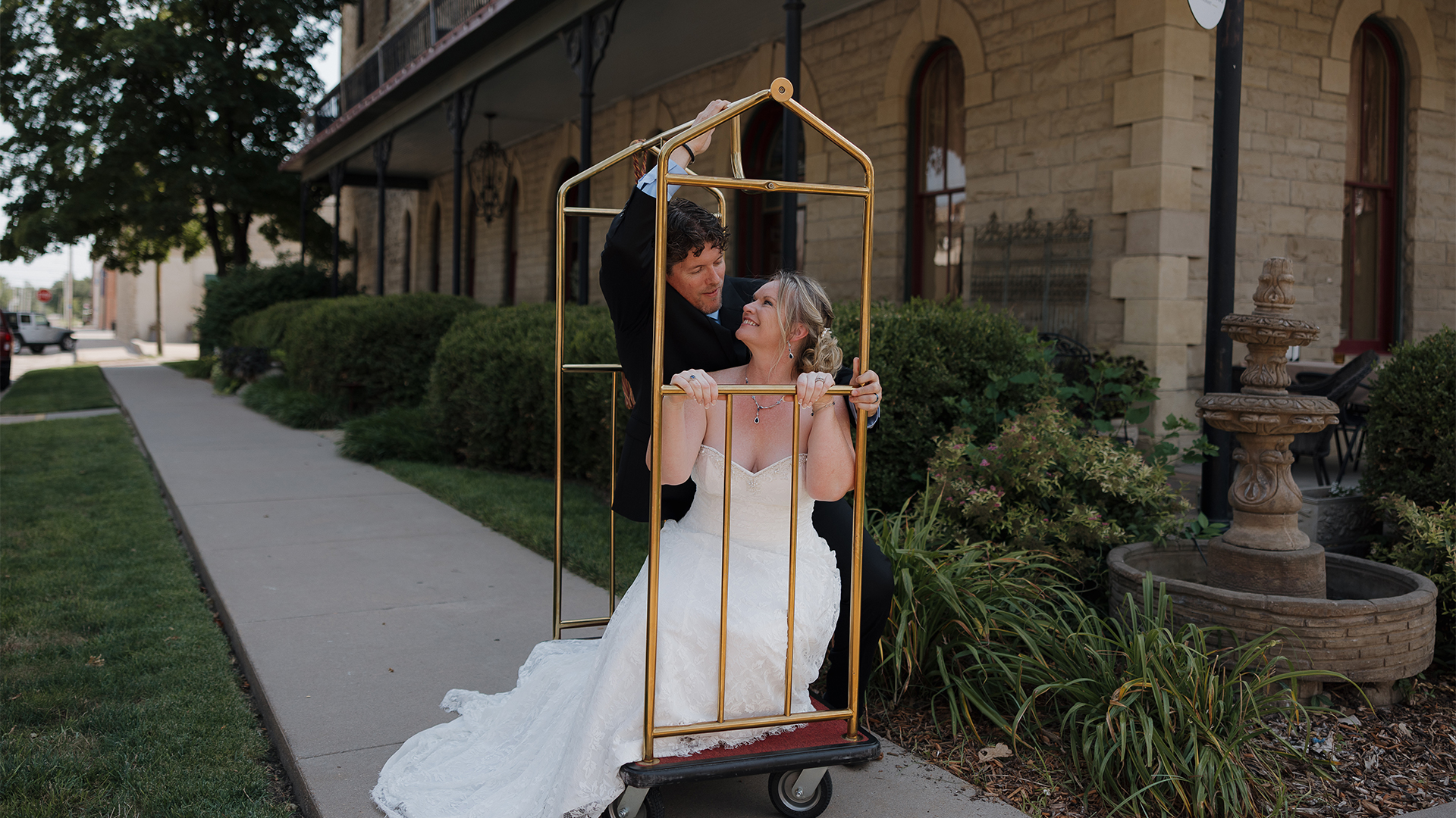 A bride and groom playfully pose inside a luggage cart outside a historic building.
