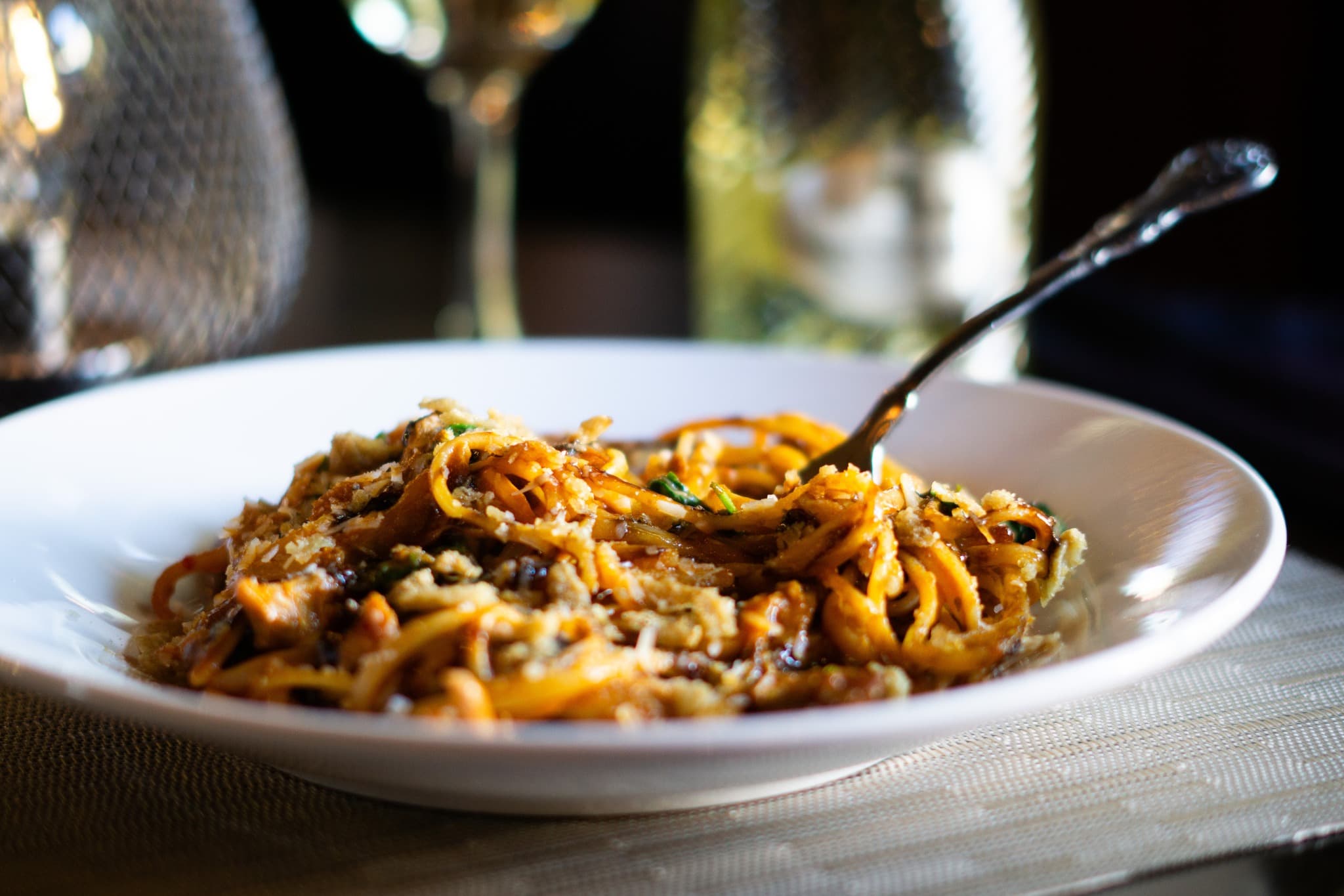 A plate of spaghetti topped with breadcrumbs and herbs, accompanied by wine glasses in the background.