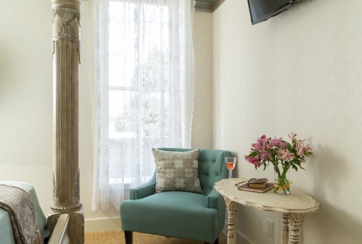 A cozy reading nook features a teal chair, a small table with flowers and a drink, and large windows with sheer curtains.