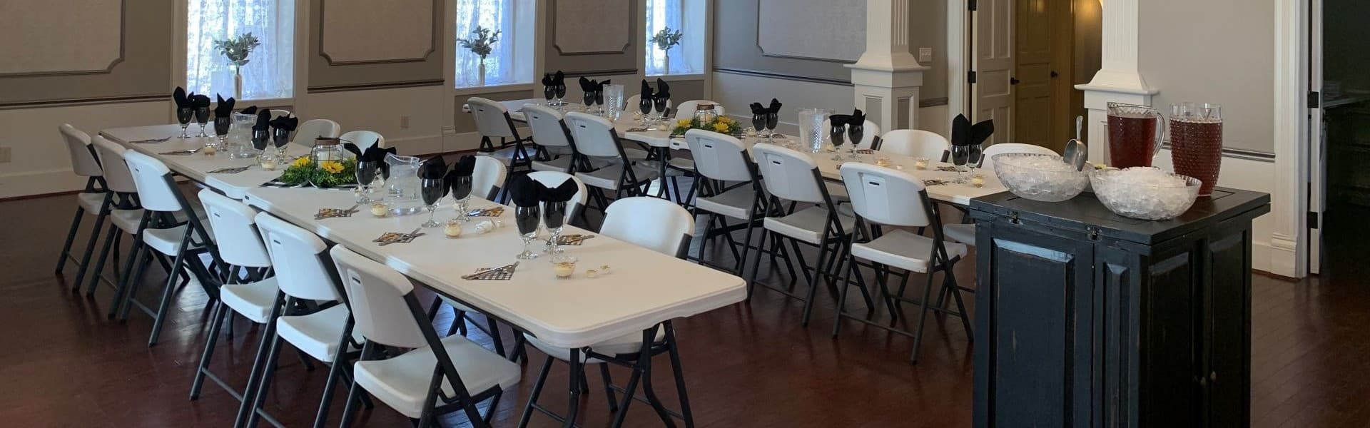 A banquet hall is set up with white folding chairs and tables, decorated with floral centerpieces and drink stations.