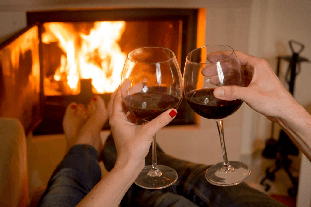 Two hands clink wine glasses in front of a cozy fireplace.