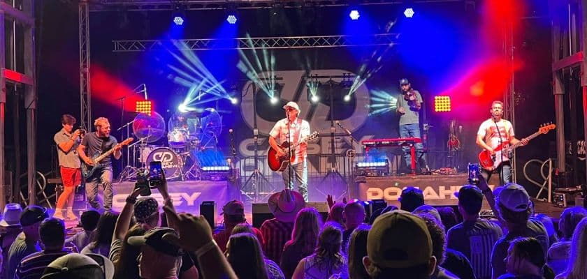 A country band performs on stage in front of a cheering crowd, with colorful lights illuminating the scene.