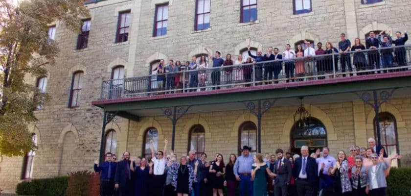 A large group of people gathers outside a historic stone building, many waving and smiling.