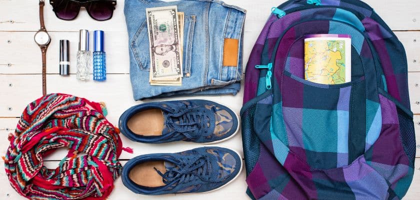 Flat lay of travel essentials including a backpack, jeans, shoes, scarf, sunglasses, watch, toiletries, and cash.
