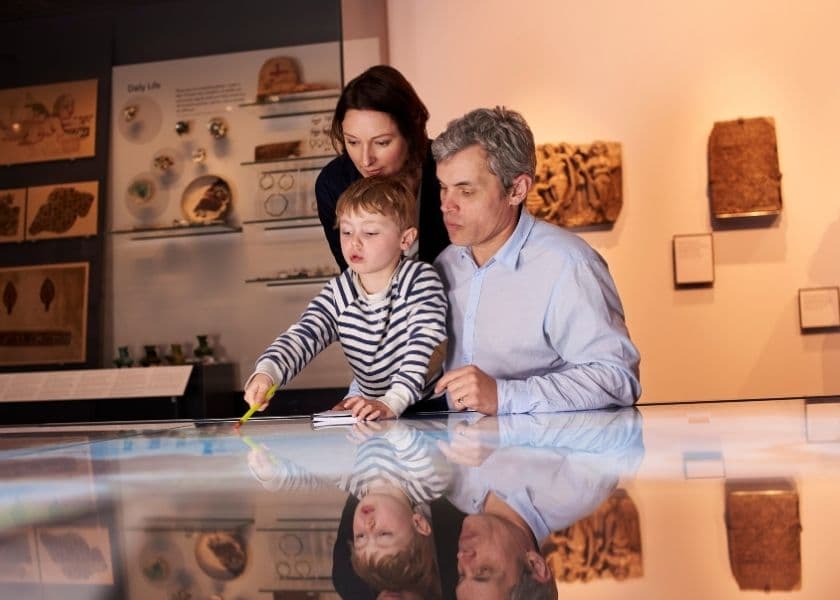 A child interacts with a touchscreen display while an adult and a parent watch in a museum setting. A child interacts with a touchscreen display while an adult and a parent watch in a museum setting.