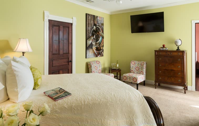 A cozy room featuring a bed, decorative chairs, a wooden dresser, and a TV on the wall with yellow walls and a piece of art.
