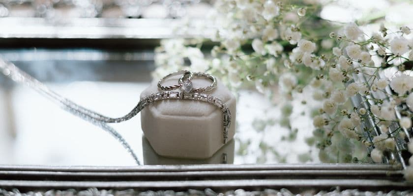 close-up of an elegant white ring box with a diamond tennis necklace with a silver baguette-cut diamonds next to a silver diamond engagement ring and matching wedding band all on a silver tray near babys breath flowers