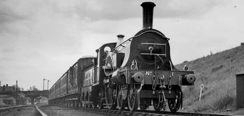 a historic black and white steam locomotive train traveling down a railway track with a bridge and small buildings in the background