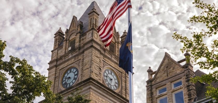 a tall stone clock tower with two large clock faces flying the american flag and another flag against a cloudy sky with some trees in the foreground