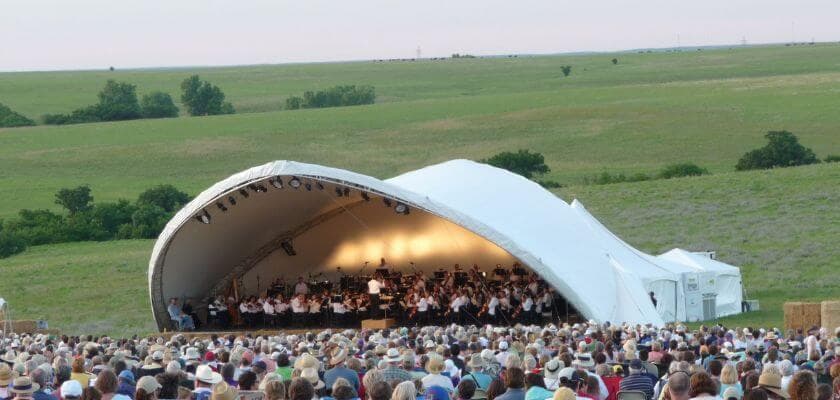 covered stage with the symphony playing in flint hills, the open prairie in the backdrop, fans seated in the foreground.