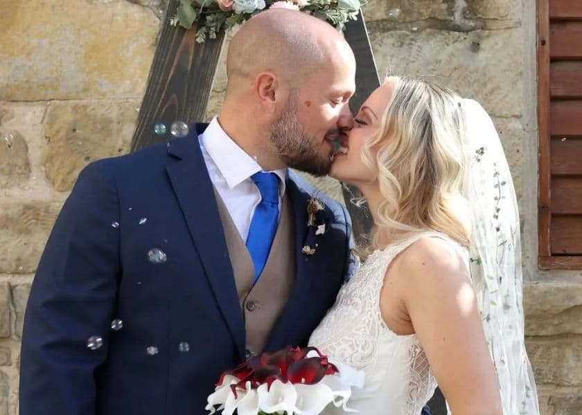 A bridal couple kissing outside under a floral arch after their elopement ceremony, while bubbles float around them.