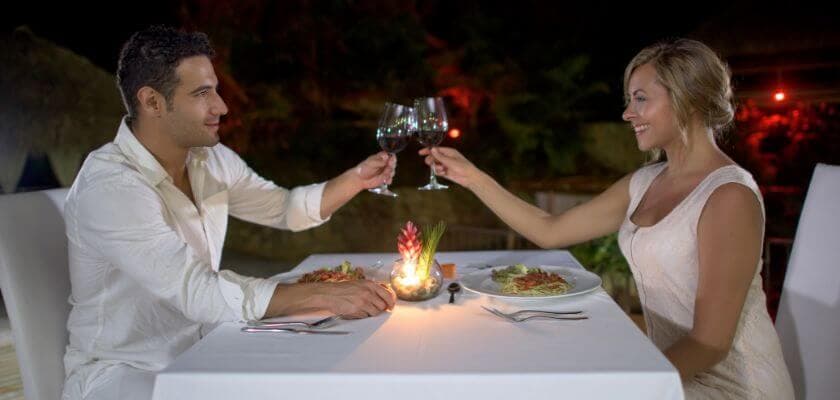 romantic couple toasting wine and dining by candlelight at a fine dining restaurant with a linen tablecloth covering the table and plants and soft ambient lighting in the backdrop